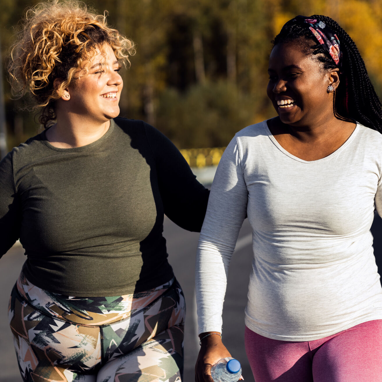 Two women wearing athletic clothing are walking outdoors, smiling and looking at each other. The woman on the left is wearing a green long-sleeve shirt and patterned leggings, and the woman on the right is wearing a light gray long-sleeve shirt and purple leggings, holding a water bottle.