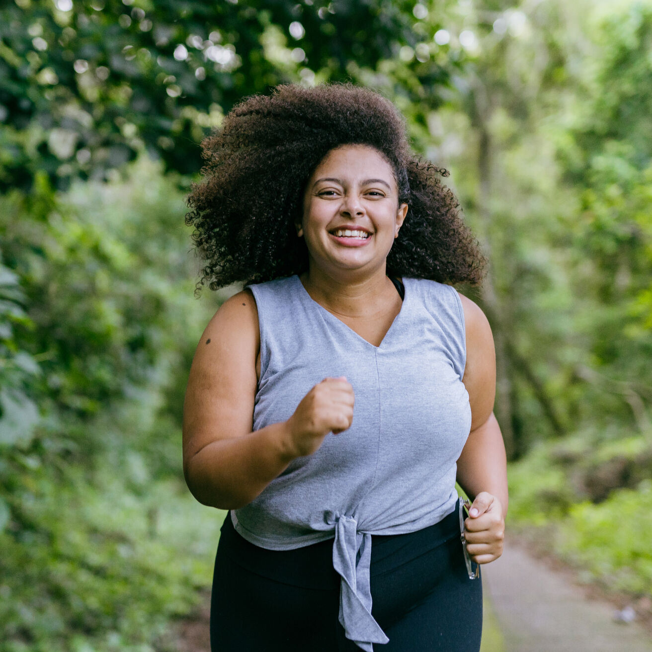 A woman with curly hair jogs on a wooded path, smiling and wearing a gray sleeveless top and black leggings. The background is lush with green trees and foliage.