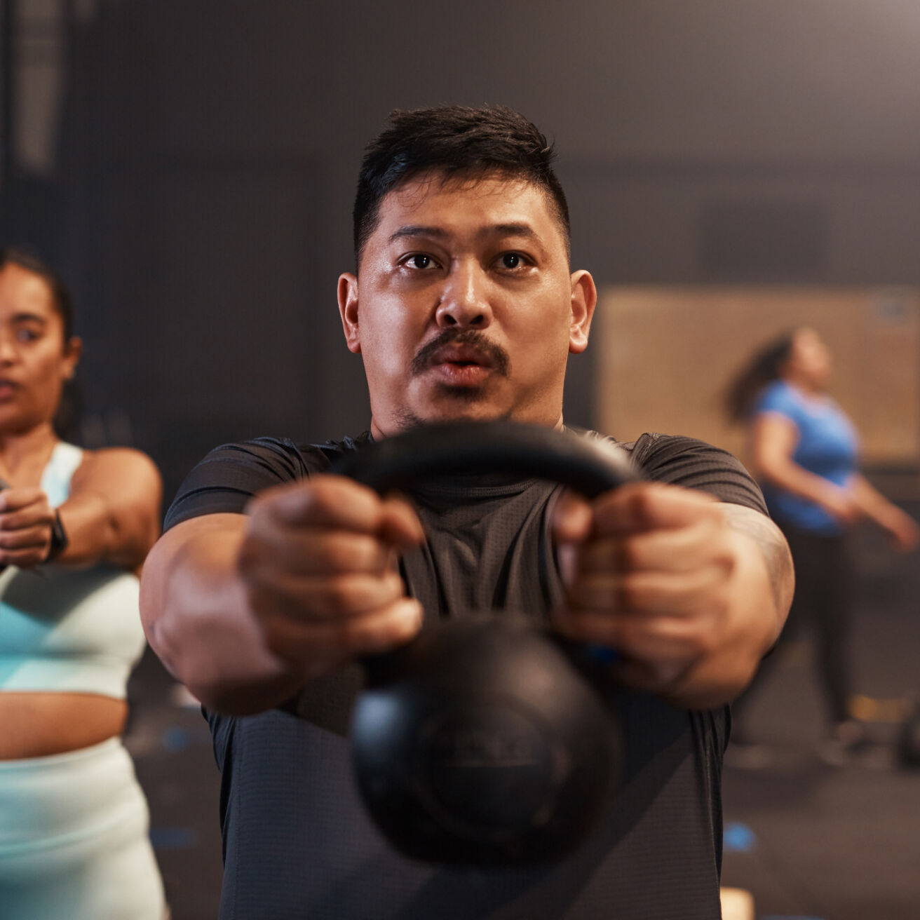 A man is performing a kettlebell workout in a gym. He is holding a kettlebell with both hands in front of him at shoulder height. Other people are working out in the background, slightly blurred.